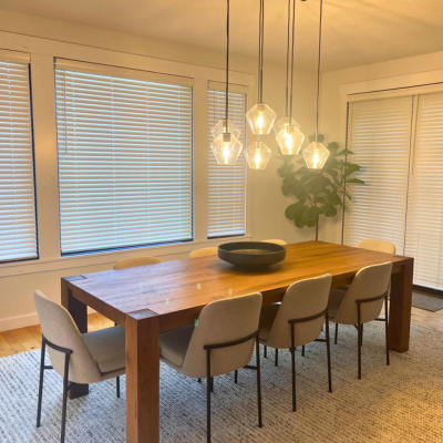A modern dining room with a large wooden table, upholstered chairs, pendant lights, and white horizontal blinds on the windows. The room features a neutral color palette with a green plant in the corner and a textured area rug on light hardwood flooring.